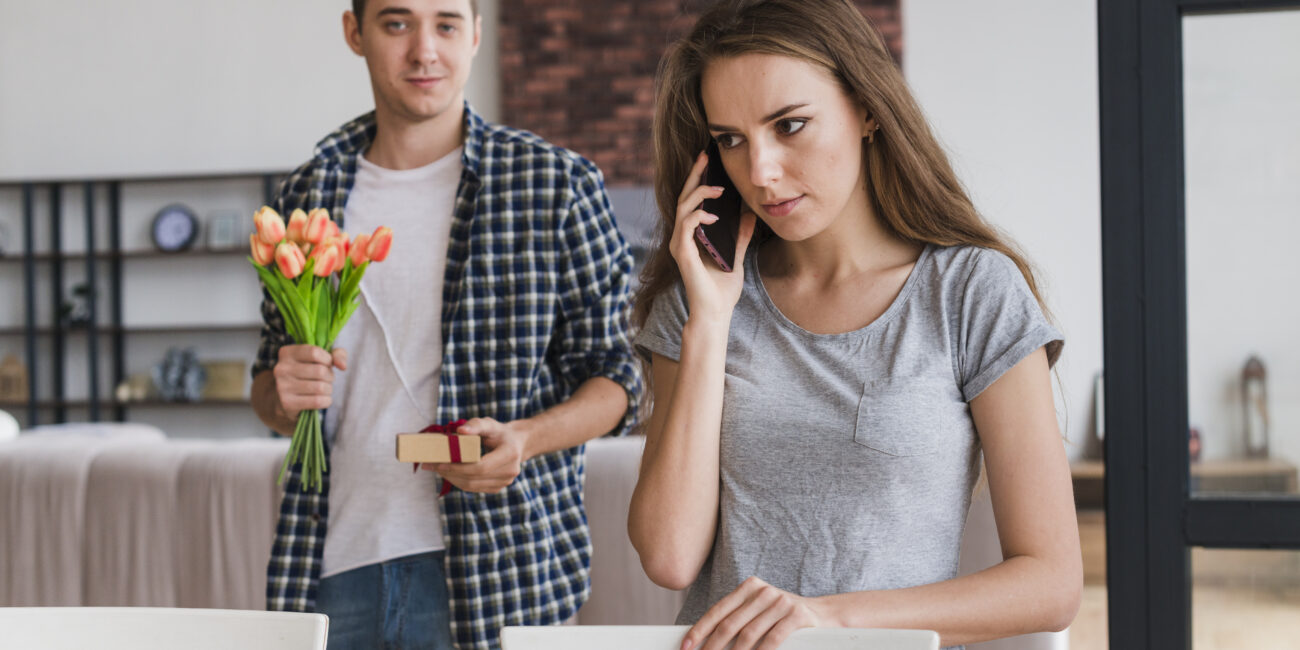 A young man holding orange tulips and a small gift box offers them to a woman on a phone call, who looks upset and distracted in a modern living room