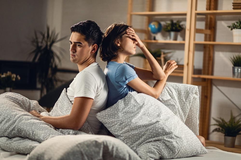 A couple sitting back-to-back in bed, appearing emotionally distant and frustrated, illustrating emotional disconnection and relationship strain over time.