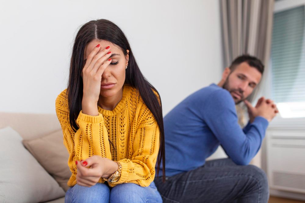 Woman sitting in emotional distress while her husband sits in the background, illustrating emotional neglect in marriage, emotional disconnection, and lack of emotional support in a relationship.